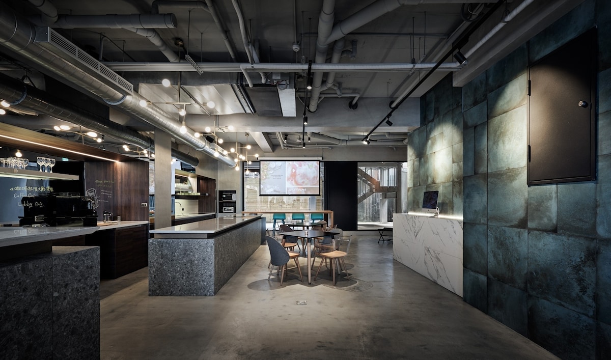 A spacious shared area combines modern design elements with industrial features, showcasing a large countertop and a dining table surrounded by wooden chairs. A large screen and contemporary decor enhance the inviting ambiance, complemented by polished concrete flooring and textured wall panels.