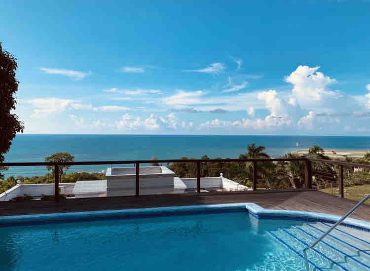 A rooftop pool glimmers under the sun, framed by a wooden deck. Beyond the pool, the expansive view reveals the Caribbean Sea with a clear blue sky and distant clouds, creating a refreshing outdoor space for relaxation.