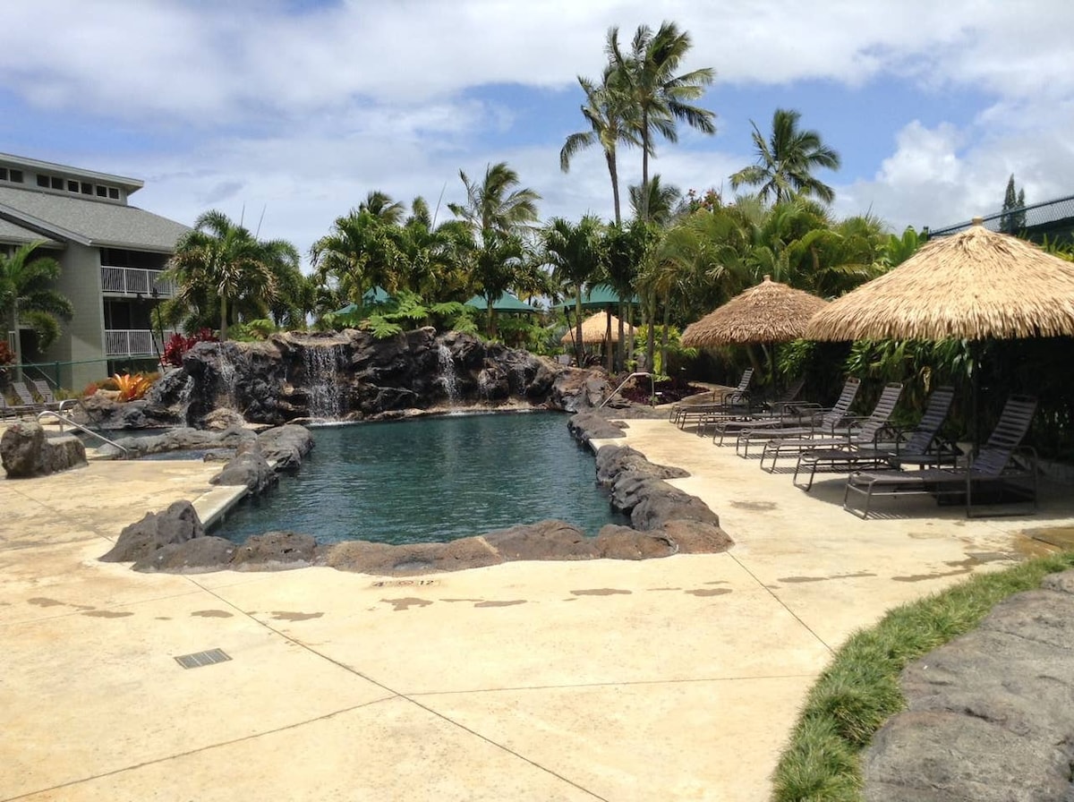 The pool area features a stunning rock waterfall set against lush tropical landscaping. Sun loungers are arranged around the pool, accompanied by thatched umbrellas providing shaded seating options. The clear water reflects the bright sky, creating a tranquil outdoor space for relaxation.