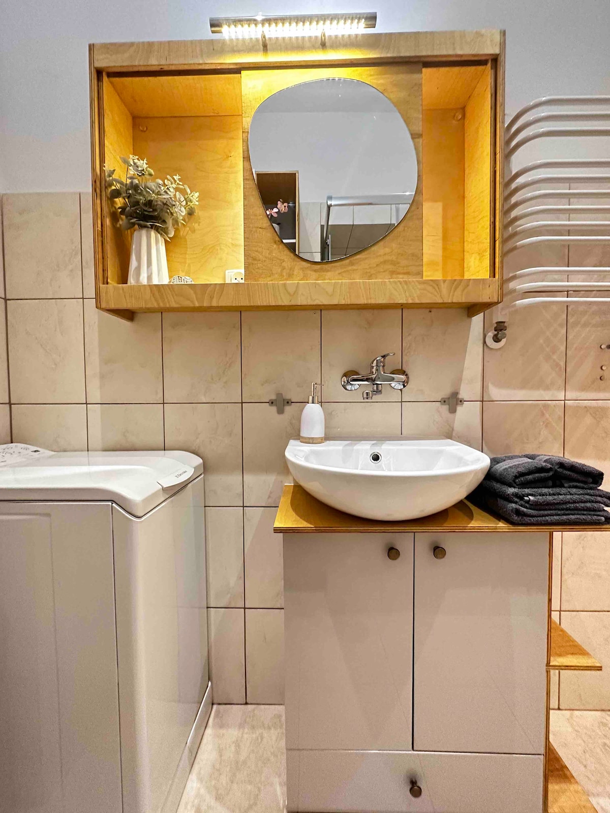 The bathroom features a modern sink with a wooden countertop and a round mirror above it. A washing machine is positioned to the left, while towels are neatly arranged on a shelf nearby. Light enhances the warm wood tones and tiled walls.