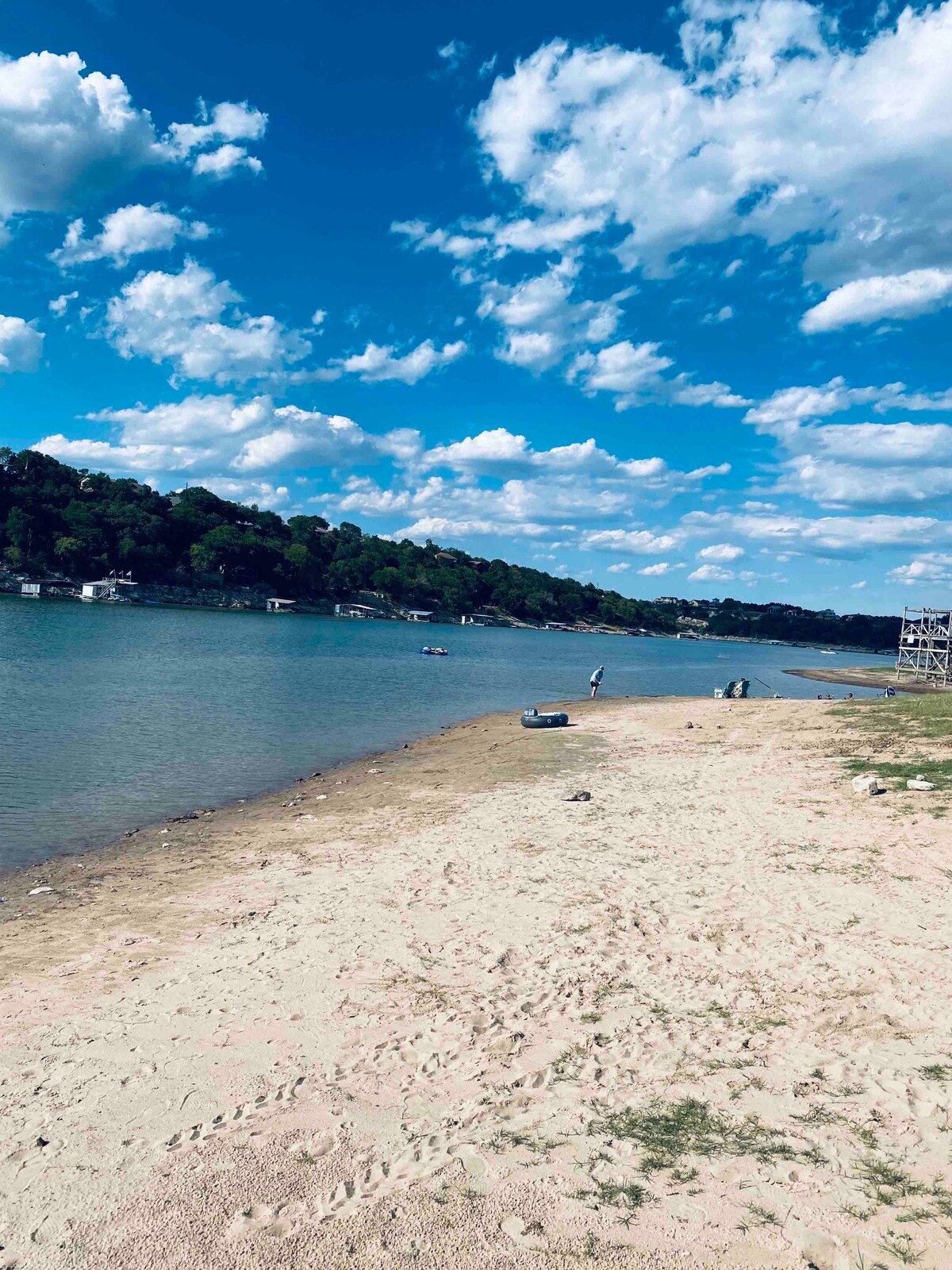 A sandy beach is visible along the edge of the Pedernales River, bordered by green trees and rolling hills in the background. The sky is bright and filled with clouds, while the calm water reflects the landscape. Small boats can be seen on the river.