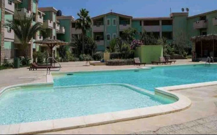 A clear swimming pool is highlighted, bordered by lounge chairs and palm trees. The surrounding area is well-maintained with lush greenery, and a shaded sitting space is visible in the background. The architecture features a light-colored facade with multiple outdoor spaces.