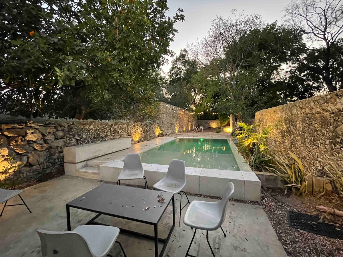 An outdoor area features a rectangular pool surrounded by natural stone walls and landscaped greenery. Four light-colored chairs are arranged around a simple table, providing a space for relaxation. Soft lighting enhances the peaceful ambiance of this tranquil setting.