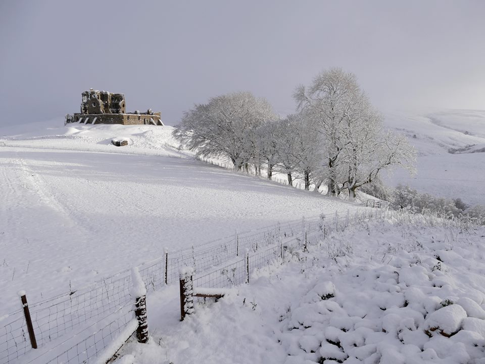 A snow-covered landscape features the ruins of Auchindoun Castle, perched atop a hill. Trees, coated in frost, line the foreground, while a gentle slope leads down to a boundary fence. The scene reflects a serene winter atmosphere, with soft, white snow covering the ground.