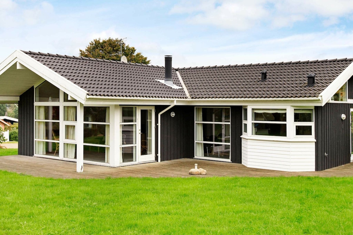 A modern holiday home is showcased with a blend of dark and light siding, featuring large windows that allow natural light to fill the interior. The outdoor space is framed by a well-maintained green lawn, contributing to a welcoming and open atmosphere.