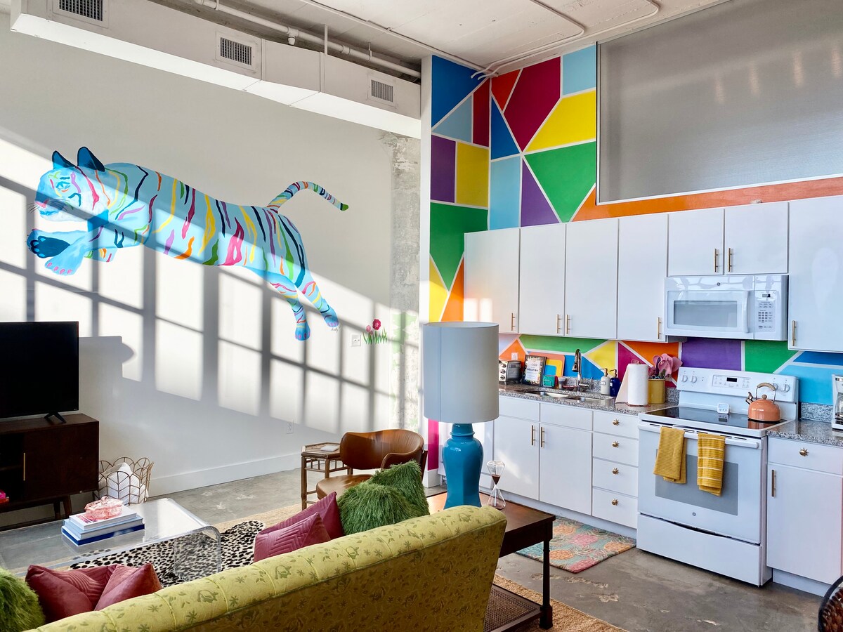 A vibrant living space features a playful mural of a colorful cat along one wall, complemented by geometric shapes in bright hues. White cabinetry houses kitchen appliances, while natural light filters through a large window, casting shadows across the polished concrete floor.