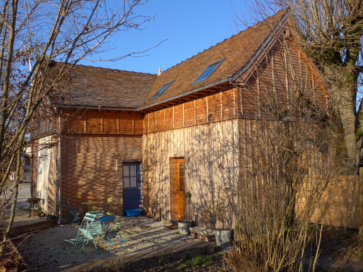 The exterior of a charming house is depicted, showcasing a blend of wood and stone materials. The structure features a distinctive gabled roof with skylights. A small outdoor seating area with blue chairs is visible, surrounded by gravel and greenery.