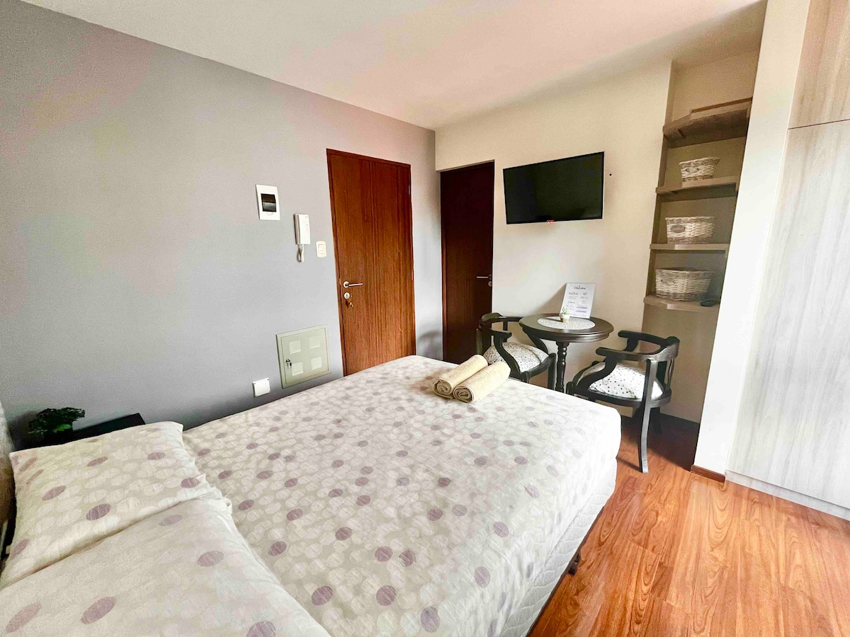 A cozy bedroom is equipped with a spacious bed featuring a patterned comforter. A small table with two chairs is positioned beside it. A wall-mounted television is visible, and storage shelves are seen in the corner, providing practical space for belongings.