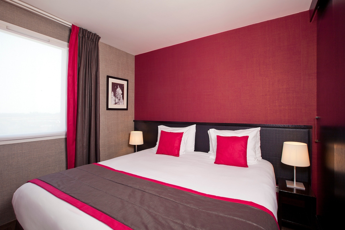 A sleeping area is presented with a spacious bed, featuring white linens and red accent pillows. Soft bedside lamps provide gentle illumination. The walls are decorated with a combination of warm red and neutral tones, complemented by patterned curtains and an artwork piece on display.