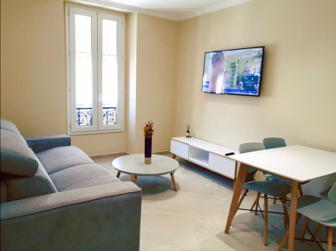 A modern living area features a light grey sofa facing a mounted television. A white coffee table rests on the floor, complemented by a small bouquet of flowers. A dining table with four blue chairs is positioned nearby, while two large windows allow ample natural light.