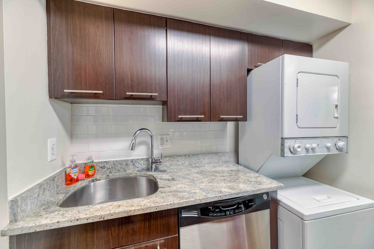 A modern kitchen space features dark wooden cabinetry complemented by a granite countertop. A stainless steel sink is positioned under a wall-mounted cabinet. To the right, a stacked washer and dryer unit is visible, enhancing the apartment's functionality.