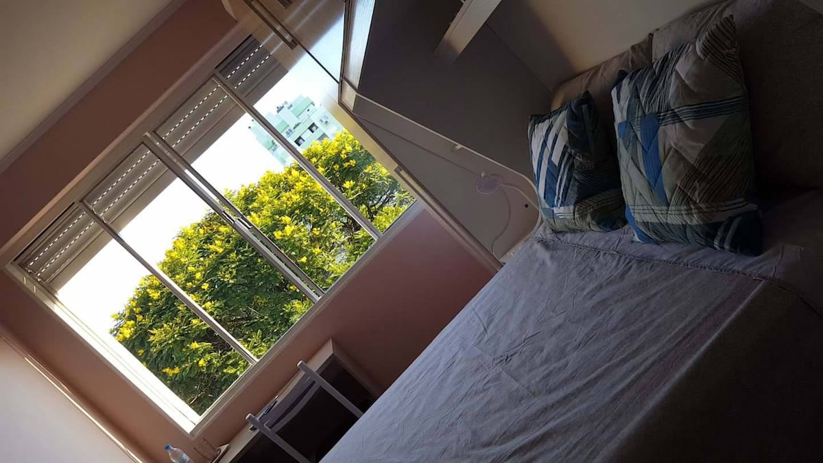 A bright sleeping area features a spacious bed adorned with patterned pillows. Large windows allow natural light to fill the room, providing a view of lush greenery outside. A simple chair and small desk are positioned nearby, creating a comfortable nook.
