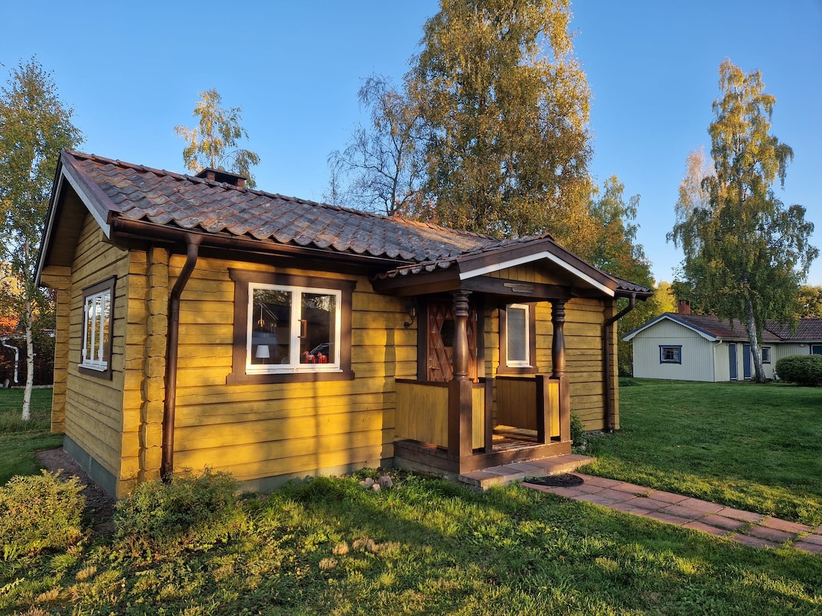 The exterior of the yellow main house features a warm wooden facade with a tiled roof. A covered porch welcomes guests, surrounded by a lush green lawn and trees. The charming architecture showcases a blend of rustic style and modern comfort.