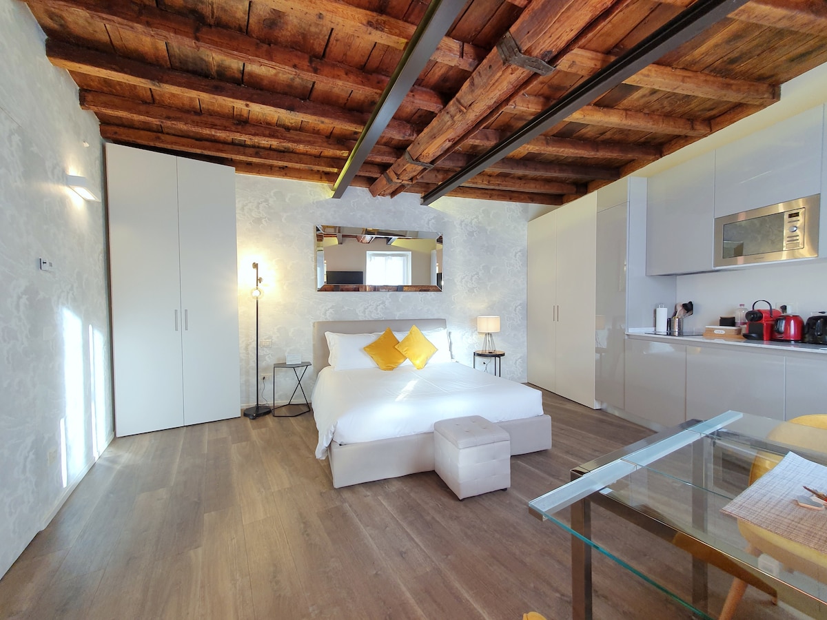 A spacious room is furnished with a large bed adorned with yellow accent pillows. A glass table sits in front of the bed, and built-in cabinets provide storage. The ceiling features exposed wooden beams, while light from the window illuminates the neutral-toned walls.