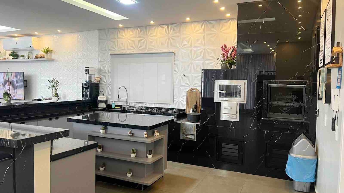 A modern kitchen displays a sleek design with black and white surfaces. The space features ample counter space, contemporary appliances, and an island at the center with decorative plants. Soft lighting highlights the textured wall and enhances the room's functionality.