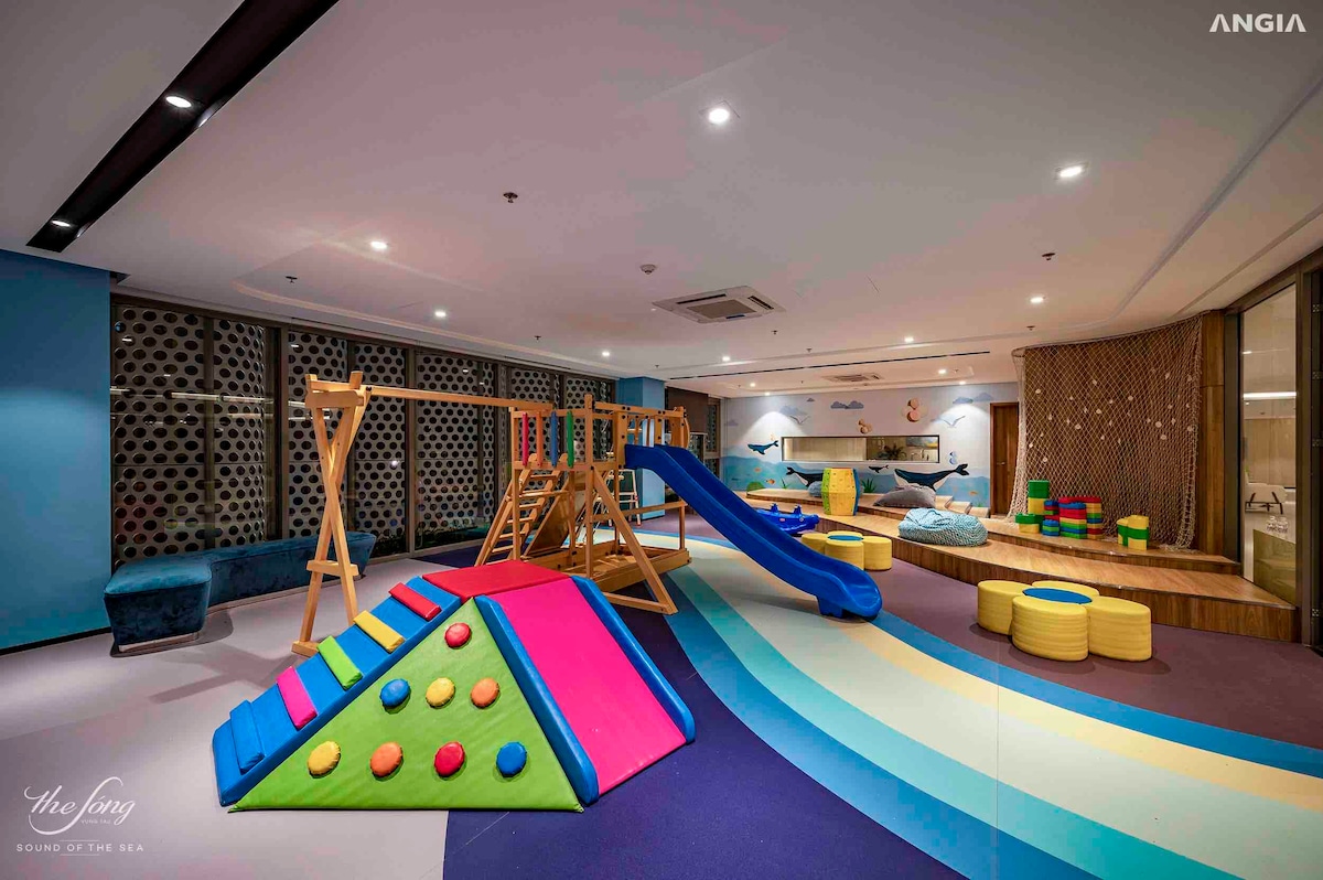 A colorful play area features a wooden climbing structure, a slide, and a soft play pyramid with bright shapes for climbing. Soft seating cushions offer a comfortable space for relaxation, while the walls are adorned with playful decorations, creating a welcoming environment for children.