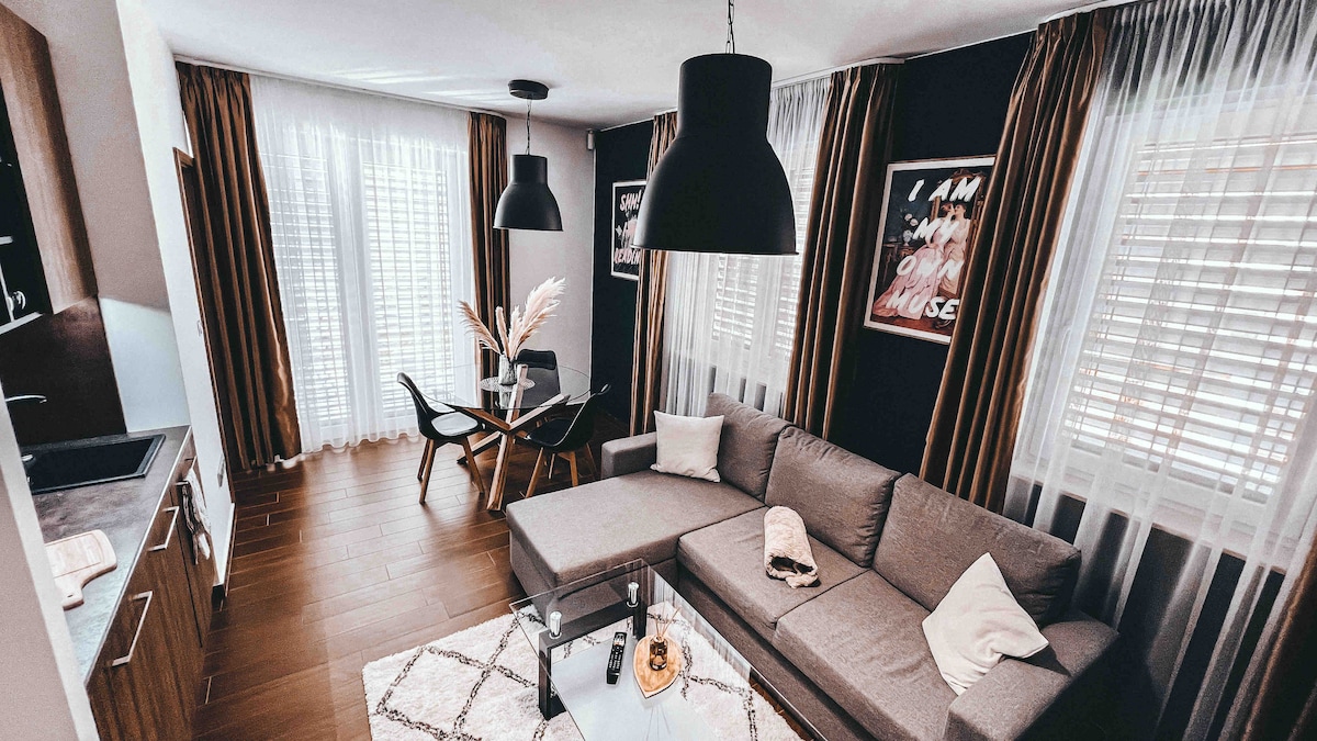 The living area features a comfortable gray sectional sofa, complemented by a glass coffee table. A dining table with chairs is positioned nearby, and large windows with sheer curtains allow natural light to fill the space. Decorative elements and modern lighting enhance the inviting atmosphere.