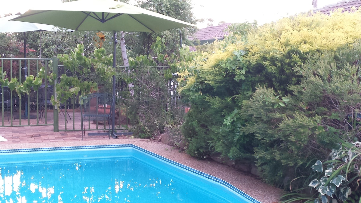A serene outdoor area surrounding a swimming pool is showcased, featuring lush greenery and well-maintained plants. A large umbrella provides shade over a seating area, enhancing the tranquility of the space. The pool's clear water reflects the surrounding foliage.