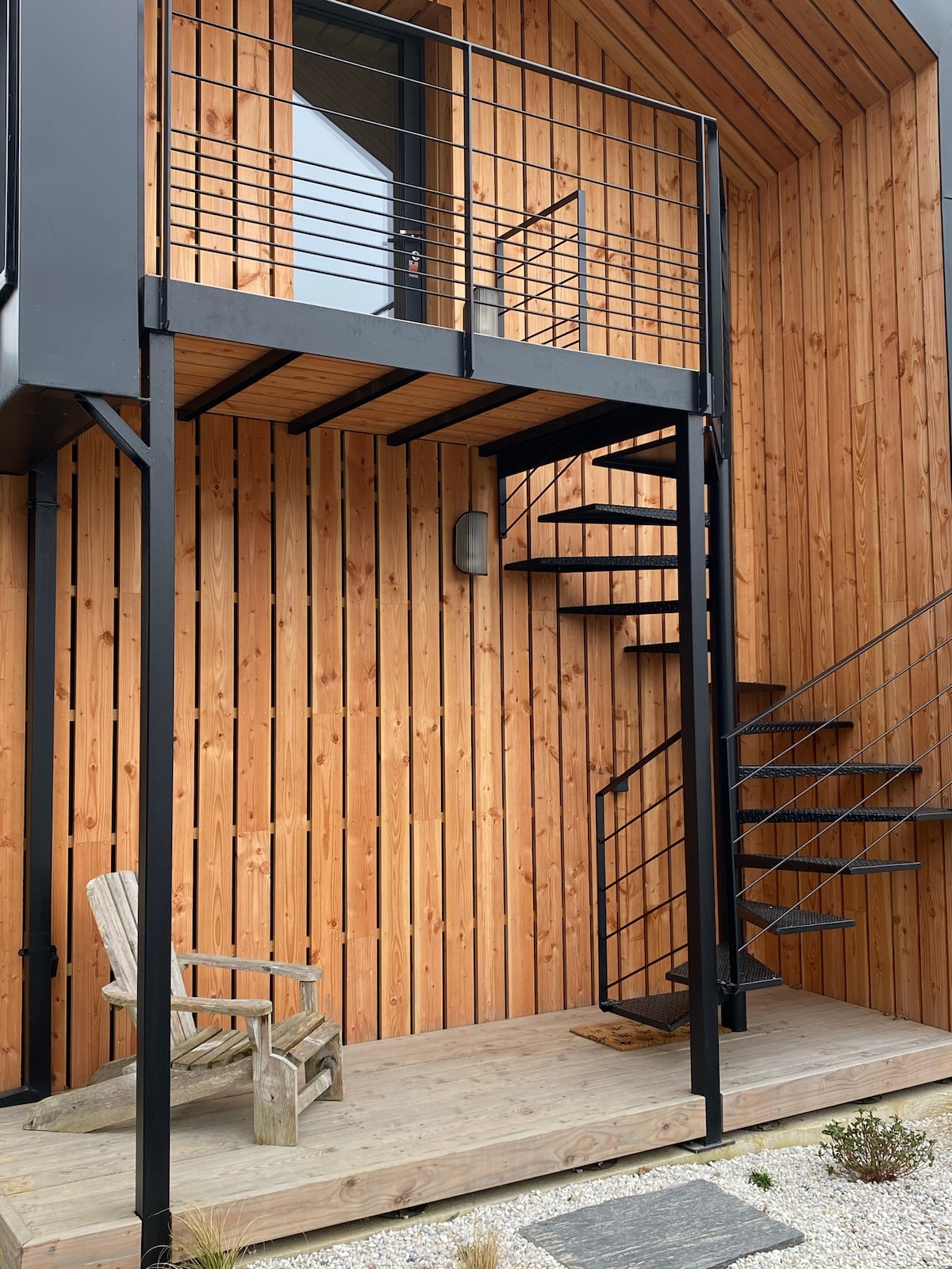A wooden exterior showcases a modern loft with a minimalist balcony, supported by a black metal frame. A spiral staircase leads to the upper level, while a wooden deck features a simple chair. The warm tones of the wooden slats are complemented by a gravel area.