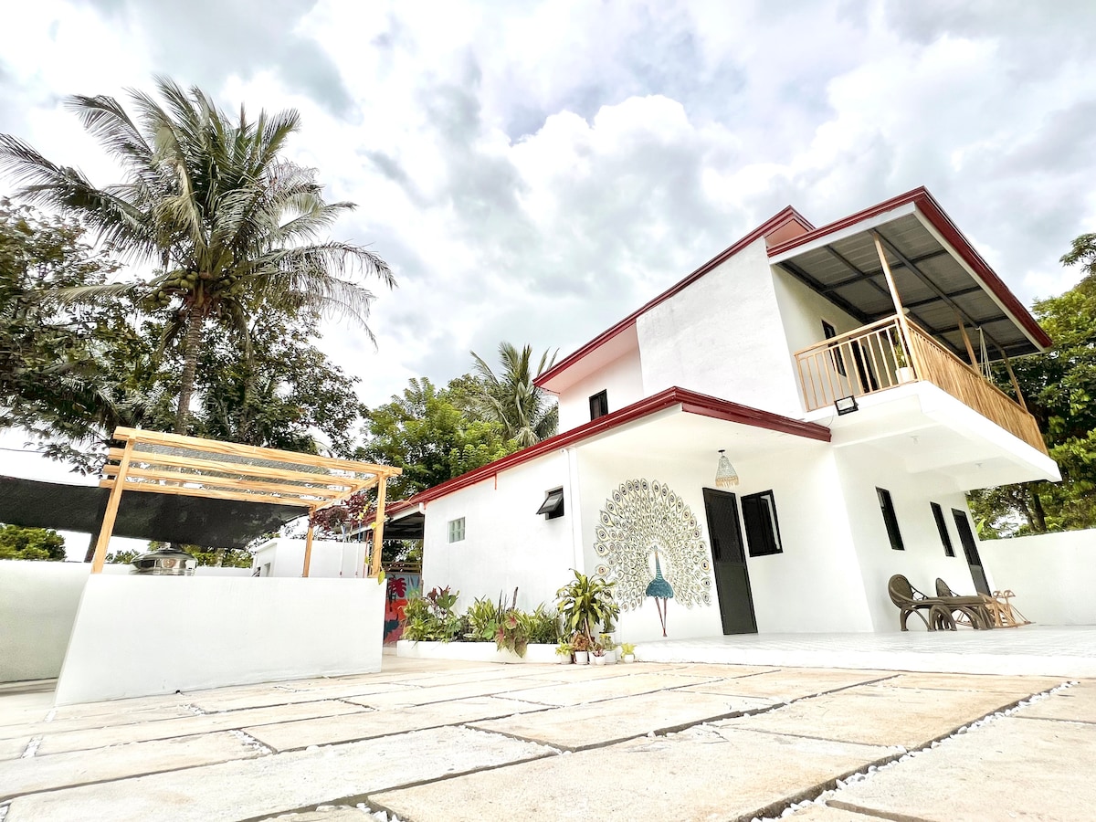 The exterior of the villa showcases a modern design with clean lines and a vibrant mural on the wall. A spacious patio area is evident, surrounded by lush greenery and palm trees. Comfortable seating arrangements provide a welcoming space for relaxation.