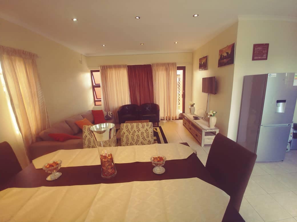 A bright living and dining area is presented, featuring a light-colored dining table with decorative bowls and a cozy seating arrangement with sofas. Natural light filters through sheer curtains, highlighting the neutral color palette and modern furnishings, including a flat-screen TV and a refrigerator.