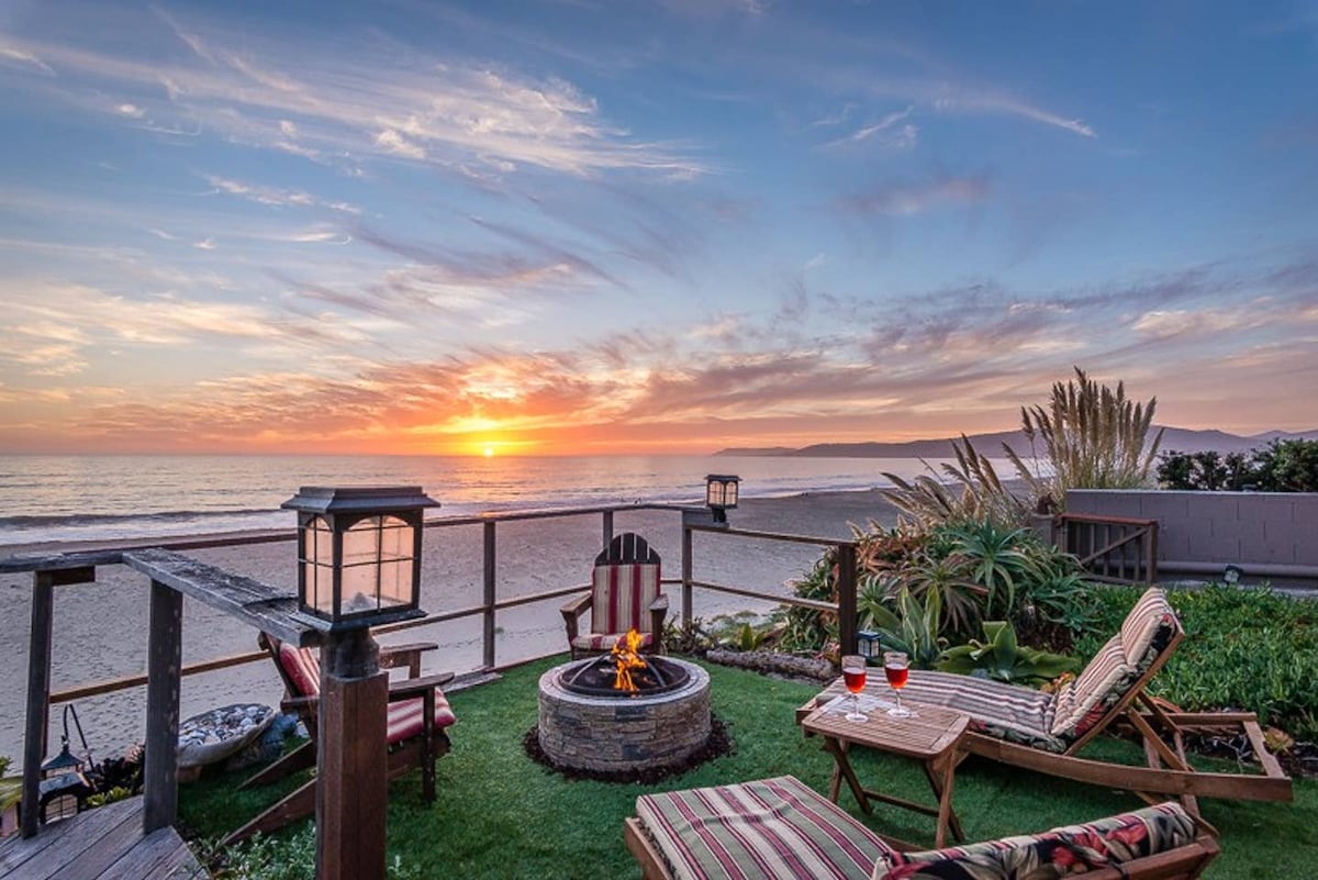 A peaceful outdoor seating area is shown with lounge chairs arranged around a fire pit. Soft lighting from lanterns enhances the ambiance as a stunning sunset is viewed over the ocean. The sound of waves can be imagined in this serene beachside setting.