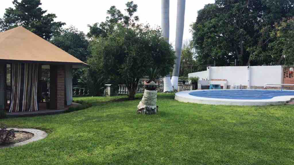 A spacious garden features lush green grass and various trees, creating a serene environment. A circular pool is visible to the right, while a palapa with a thatched roof is situated nearby, offering a shaded area for relaxation.