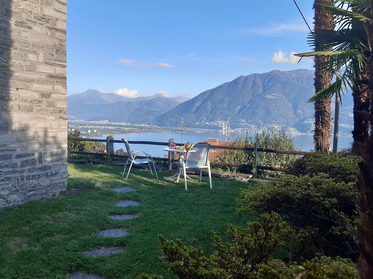 A peaceful garden area features a grassy lawn with stone pathway, leading to two outdoor chairs and a small table. Surrounding palm trees and a stone wall provide a natural backdrop, while the distant mountains and lake create a serene landscape view.