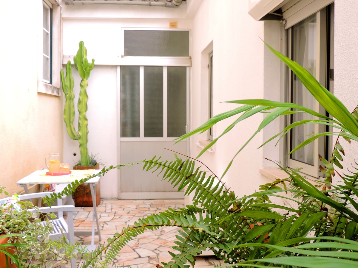 A cozy outdoor space is visible, featuring a small table set for two and surrounded by greenery. A tall cactus stands beside a glass door, while potted ferns add lushness to the scene. The area is well-lit, enhancing the tranquil environment.