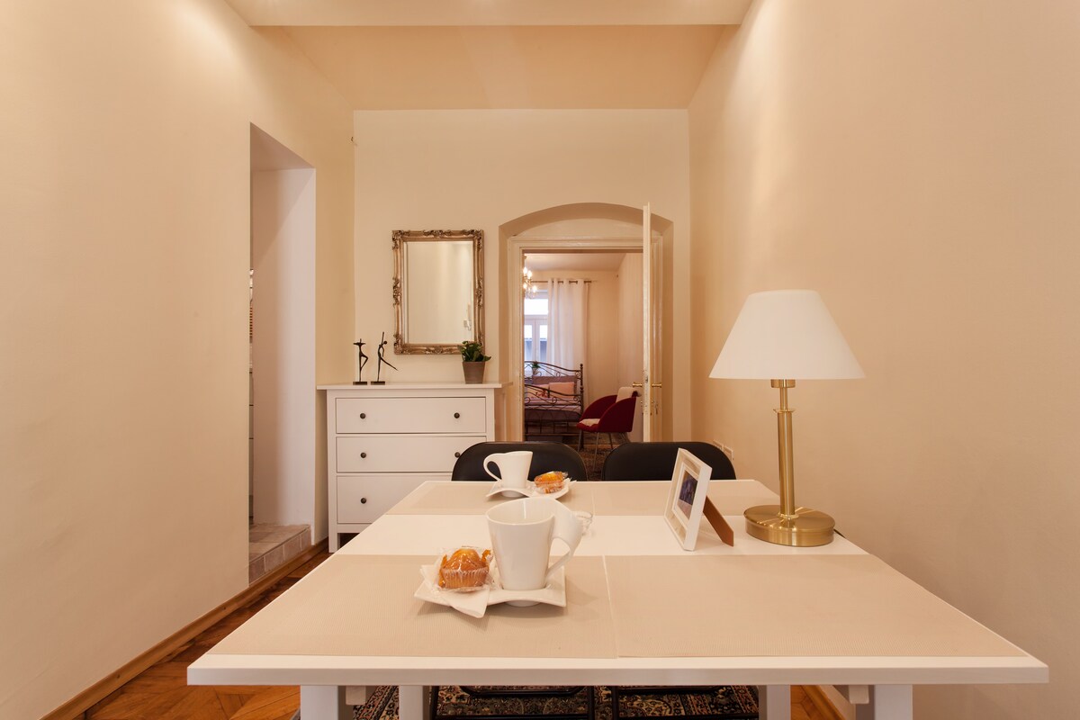 A dining area features a light table set with cups and pastries. A decorative lamp stands beside the table. The warm tones of the walls complement a white chest of drawers against the backdrop, with an adjoining archway leading to a cozy living space.