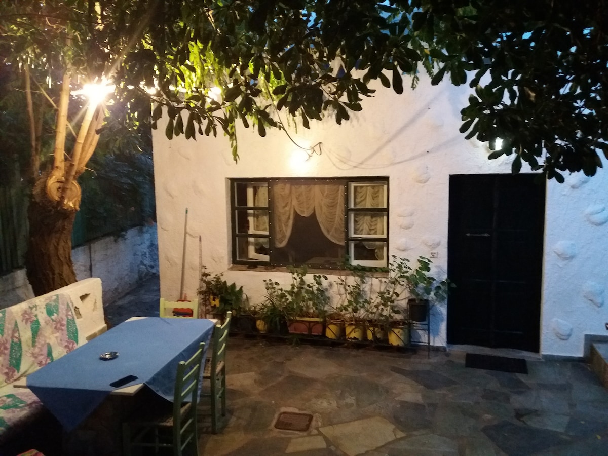 An outdoor patio area is featured, surrounded by greenery and illuminated by soft lighting. A stone floor supports a rectangular table accompanied by chairs. The white exterior of the house is partially visible, with a window adorned by sheer curtains, adding to the serene ambiance.