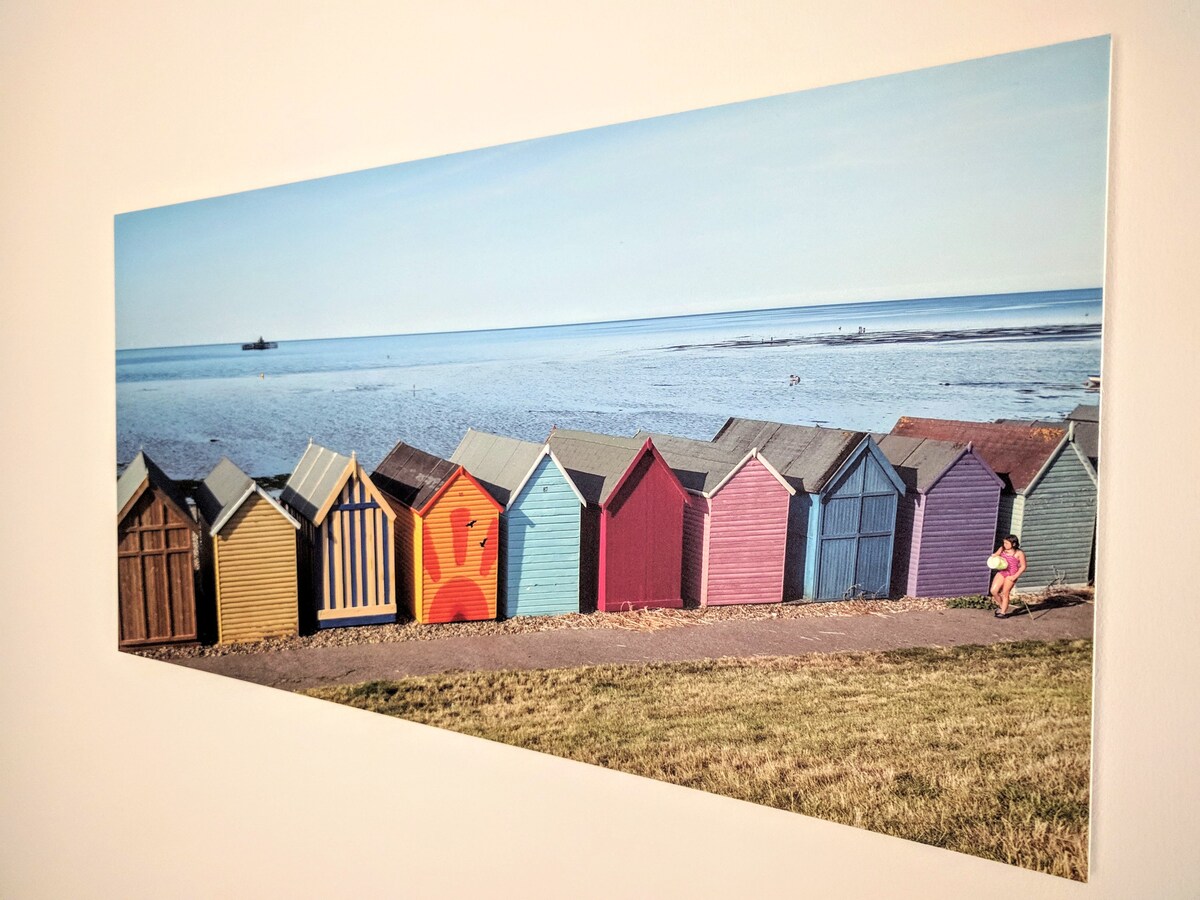 A canvas print displays a row of colorful beach huts lined along the shoreline, set against a calm sea. The horizon meets the sky in soft blue tones, while a grassy foreground adds a natural element to the scene.