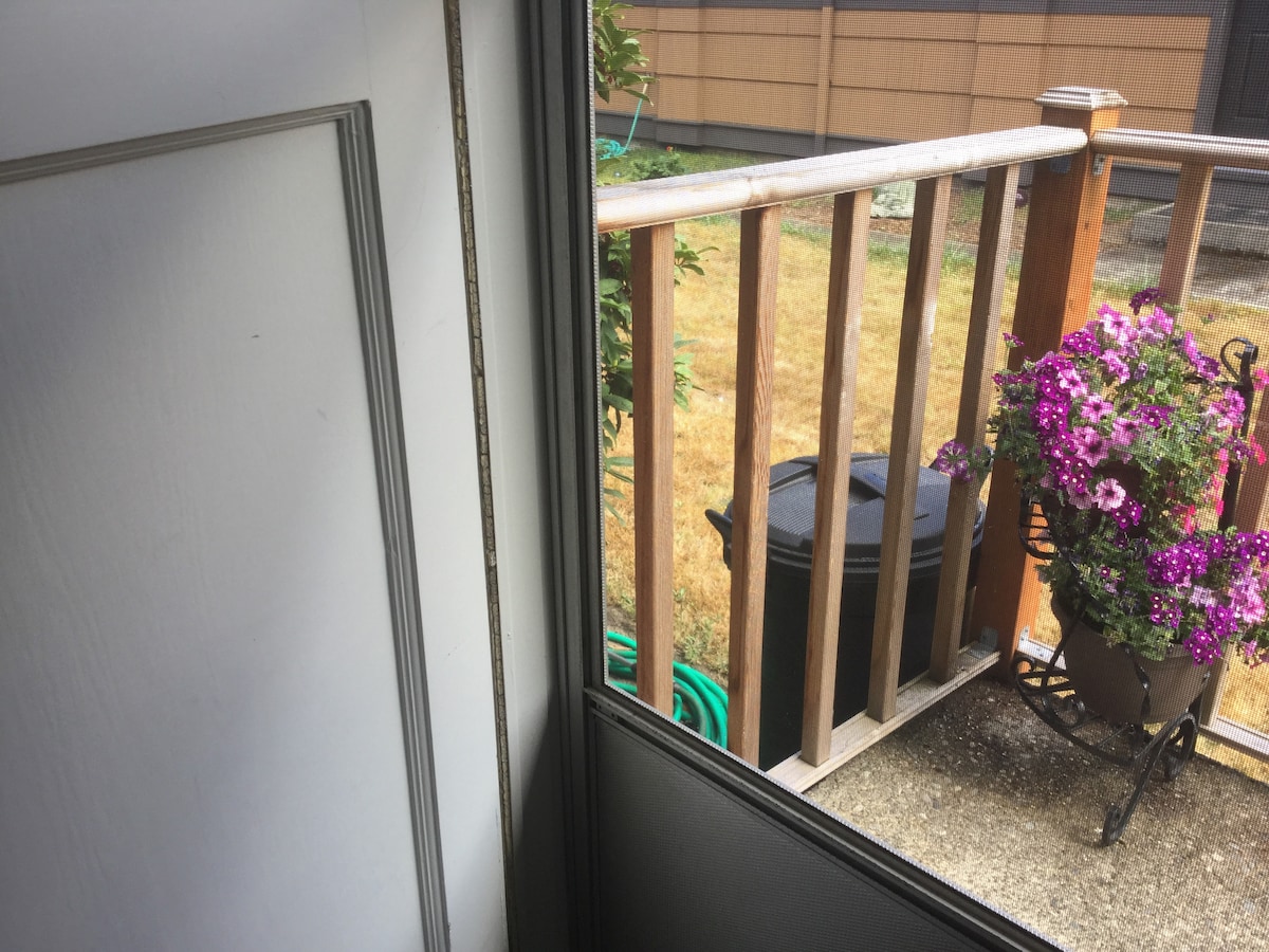 A view from the doorway to a small private porch area, featuring a wooden railing and a decorative pot filled with vibrant purple flowers. A grassy area is seen beyond, and a black container is positioned on the ground.