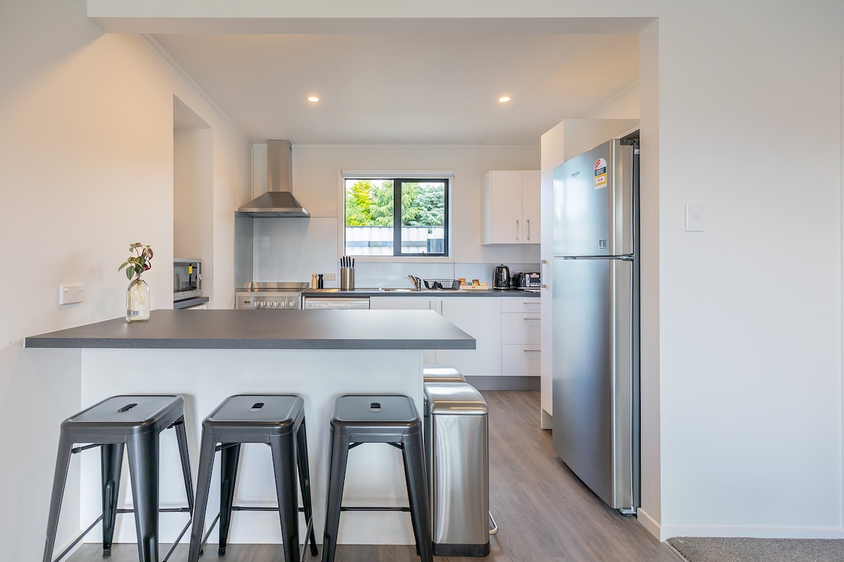 A modern kitchen is visible, featuring sleek white cabinetry and stainless steel appliances. A central island with three bar stools provides a casual dining space, while a window offers a view of the outdoors. Countertops are uncluttered, suggesting a functional cooking area.