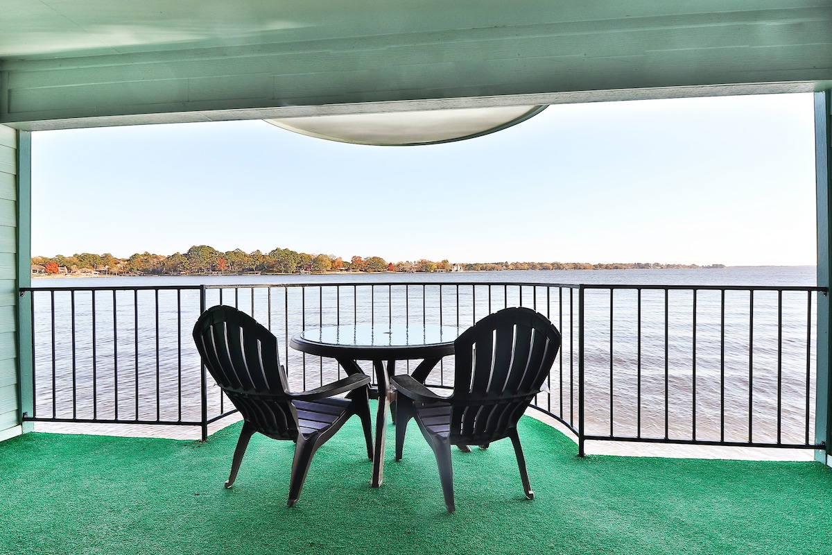 A private over-the-water balcony features two dark lounge chairs and a small round table, perfect for enjoying lake views. The railing encircles the balcony, offering an unobstructed view of the calm water and tree-lined shore in the distance.