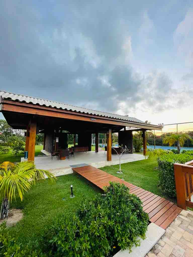 A spacious outdoor area is presented, featuring a wooden structure with a covered patio. Lush green lawns surround the space, complemented by a wooden walkway leading to the entrance. Trees and garden plants add a natural touch, enhancing the serene environment.