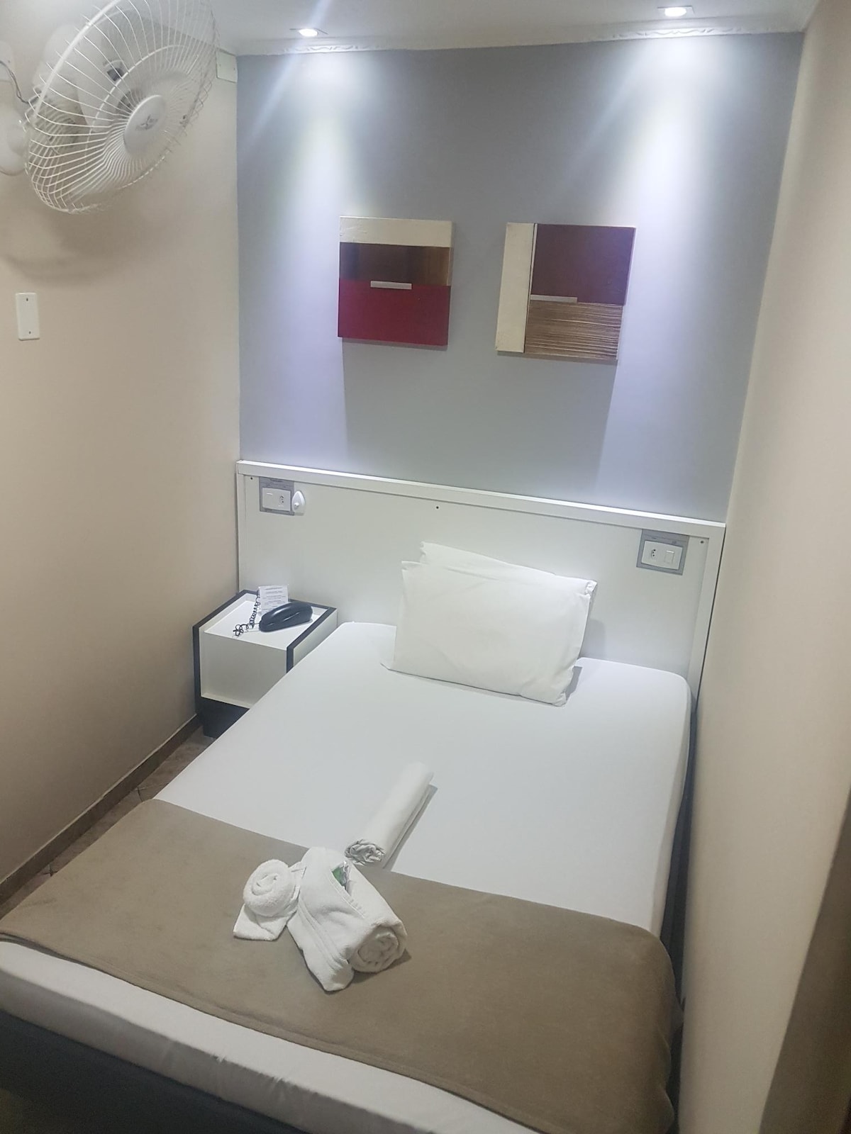 A compact sleeping area features a single bed with a white pillow and a light brown throw. The wall above is adorned with abstract artwork. A bedside table with a phone is positioned adjacent to the bed, and a ceiling fan is mounted overhead.