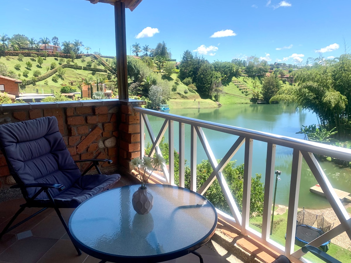 A sunlit balcony features a glass table accompanied by a comfortable chair. The scenic view includes a tranquil lake surrounded by lush greenery and gentle slopes. Clear blue skies enhance the inviting atmosphere of this outdoor space, perfect for relaxation.