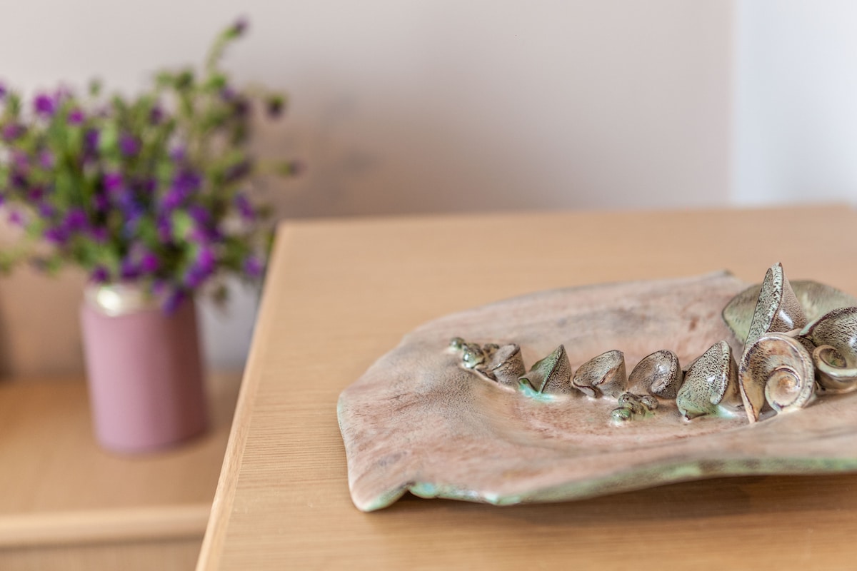 A decorative plate featuring shell motifs is displayed on a wooden surface. A small vase of purple flowers adds a touch of color in the background, enhancing the overall aesthetic of the space.