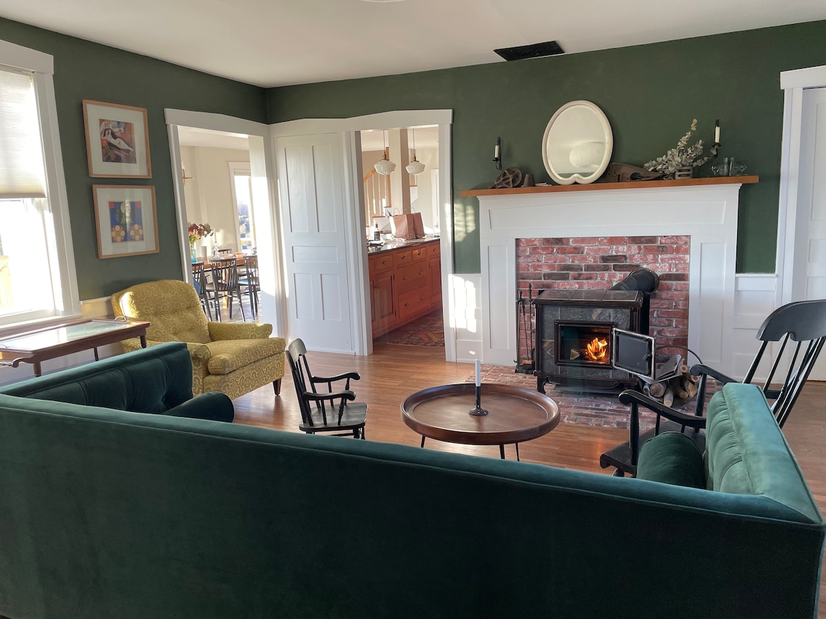 A cozy living area features a green velvet sofa and two armchairs in a warm, inviting space. A wooden coffee table sits at the center, and a brick fireplace with a small fire adds a touch of warmth. Light enters through large windows, illuminating the room.