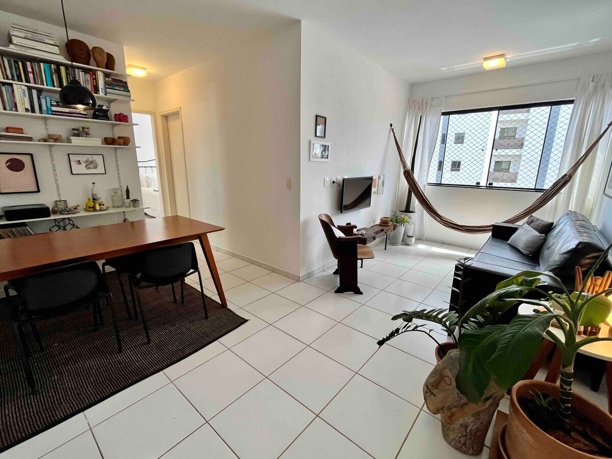 A bright and airy living space features a large window with natural light streaming in. A comfortable couch and a stylish chair are arranged around a wooden table, while a decorative plant adds a touch of greenery. A hammock is hung for relaxation.