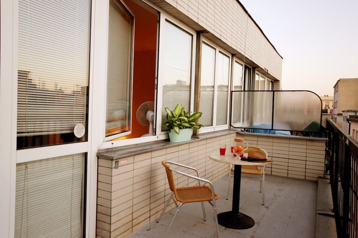 A spacious private terrace is shown, featuring a round table with two chairs. Potted greenery adds a touch of nature under the evening sky. Large windows surround the area, allowing ample natural light, while a partial view of the city is visible in the distance.