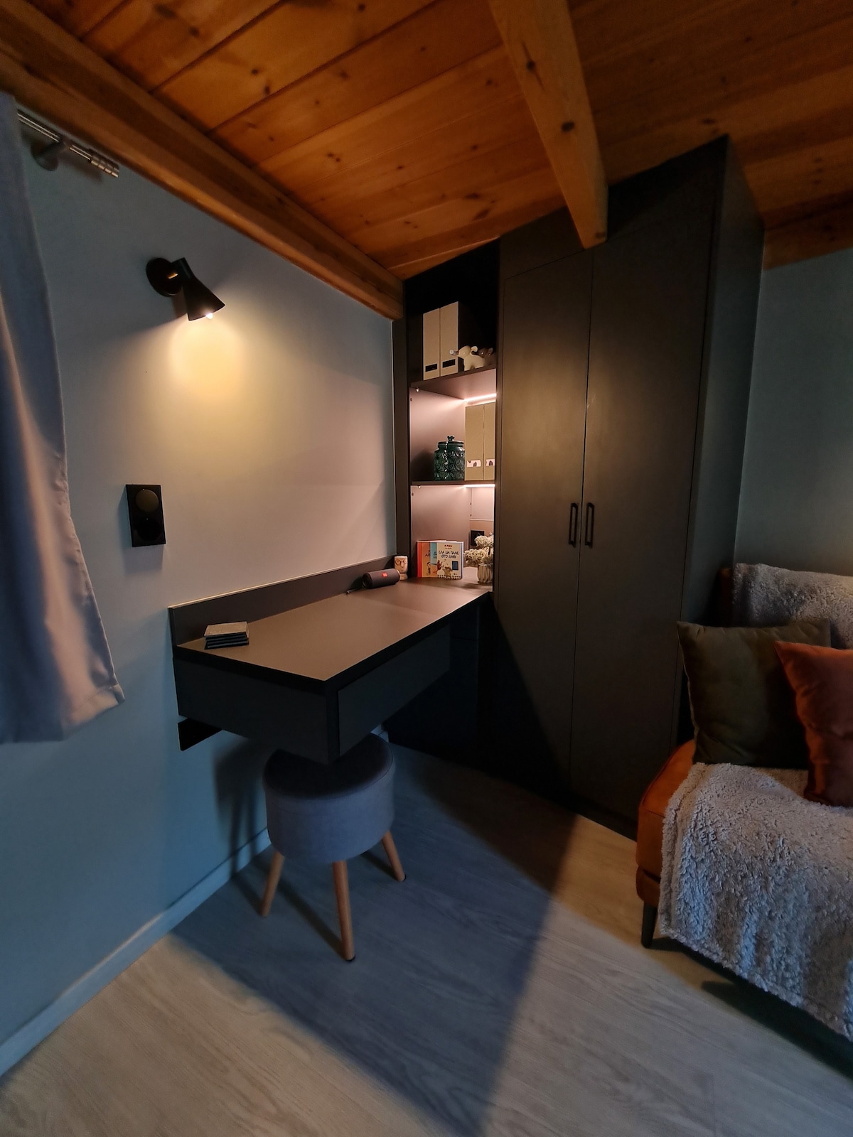 A cozy workspace is featured against a backdrop of soft blue walls, complemented by a wall-mounted light. A compact desk extends from cabinetry, while a minimalist stool provides seating. Shelving displays various items, and a plush sofa in the corner invites relaxation.