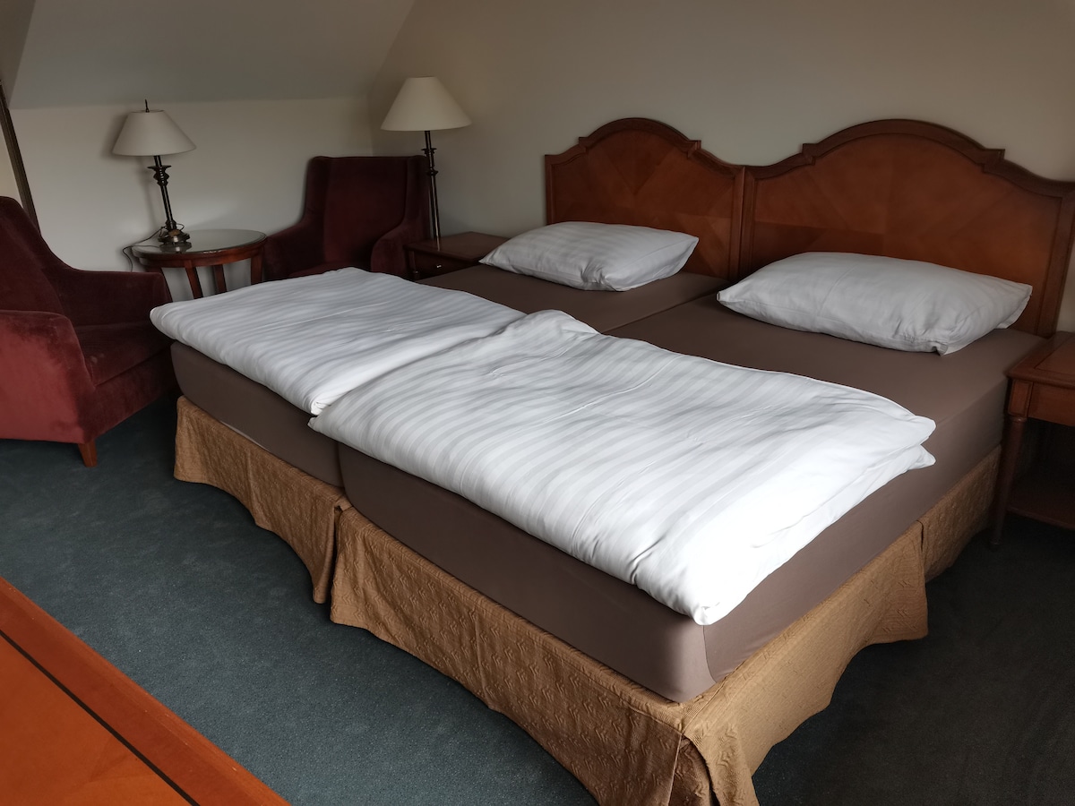 A well-appointed sleeping area features two spacious beds with light linens, positioned on a soft carpet. Flanking the beds are two armchairs in deep red upholstery and a round side table with lamps providing soft lighting.
