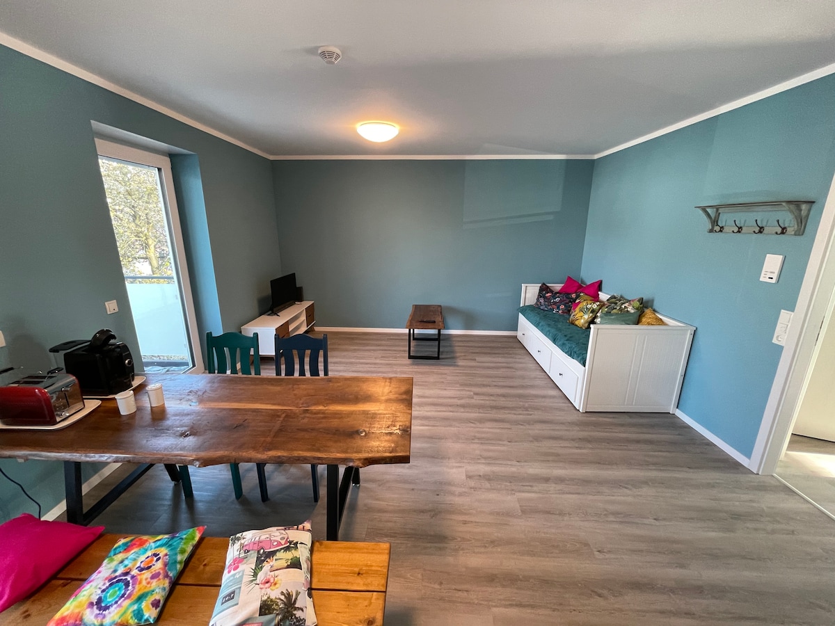 An open living area is presented with light blue walls and natural light coming from the window. A wooden dining table with colorful chairs occupies one side, while a compact seating area with decorative pillows and a coffee table is positioned on the opposite side.