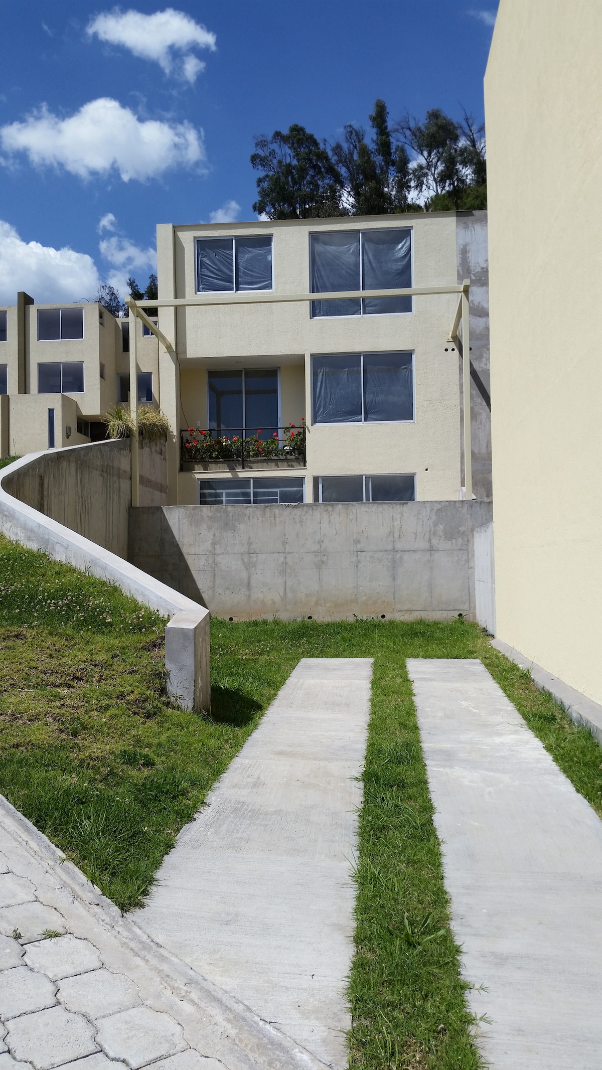 A modern two-story building is seen against a clear blue sky, featuring large windows dressed with light-colored frames. A grassy pathway leads to the entrance, flanked by concrete walkways that provide accessibility. Flower boxes are visible on the upper level, adding a touch of greenery.