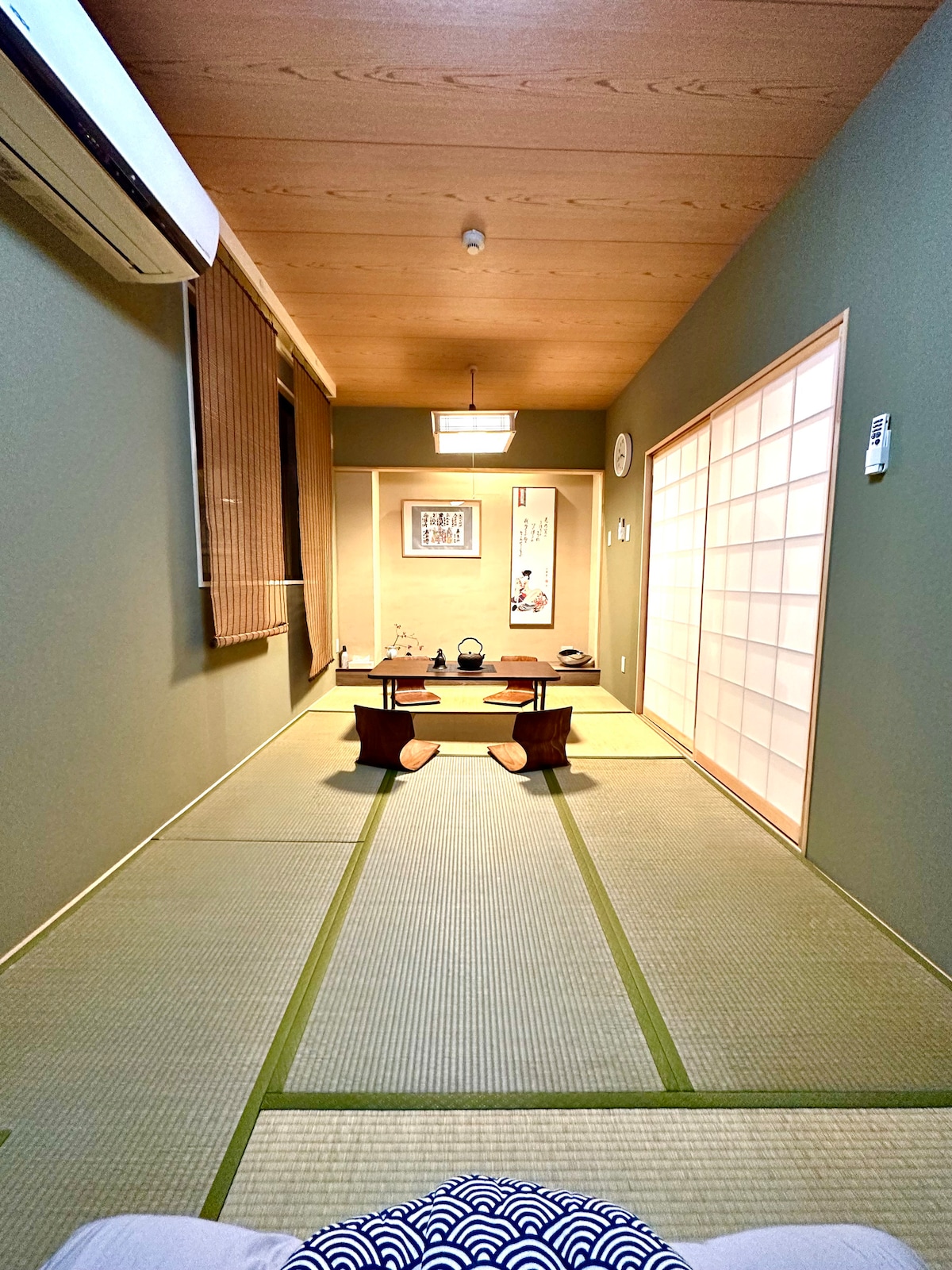 A traditional Japanese room is showcased with tatami mat flooring and wooden accents. A low wooden table is positioned at the center, surrounded by seating. Shoji screens allow natural light, while green walls contribute to a calming ambiance. Decorative elements are present in the background.