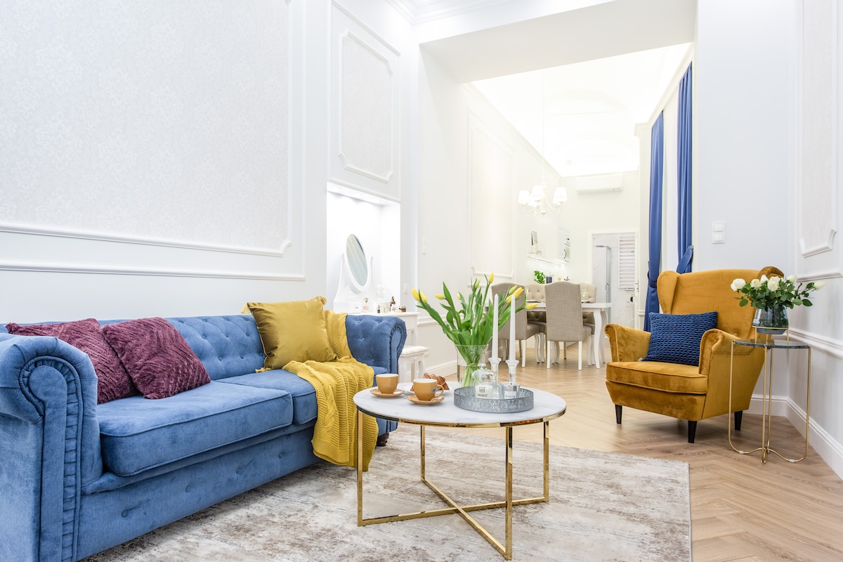 A spacious living area features a blue upholstered sofa, complemented by decorative cushions and a throw. A round coffee table sits at the center, adorned with a tray containing two cups and a small vase. Soft lighting highlights the elegant decor and adjoining dining space.