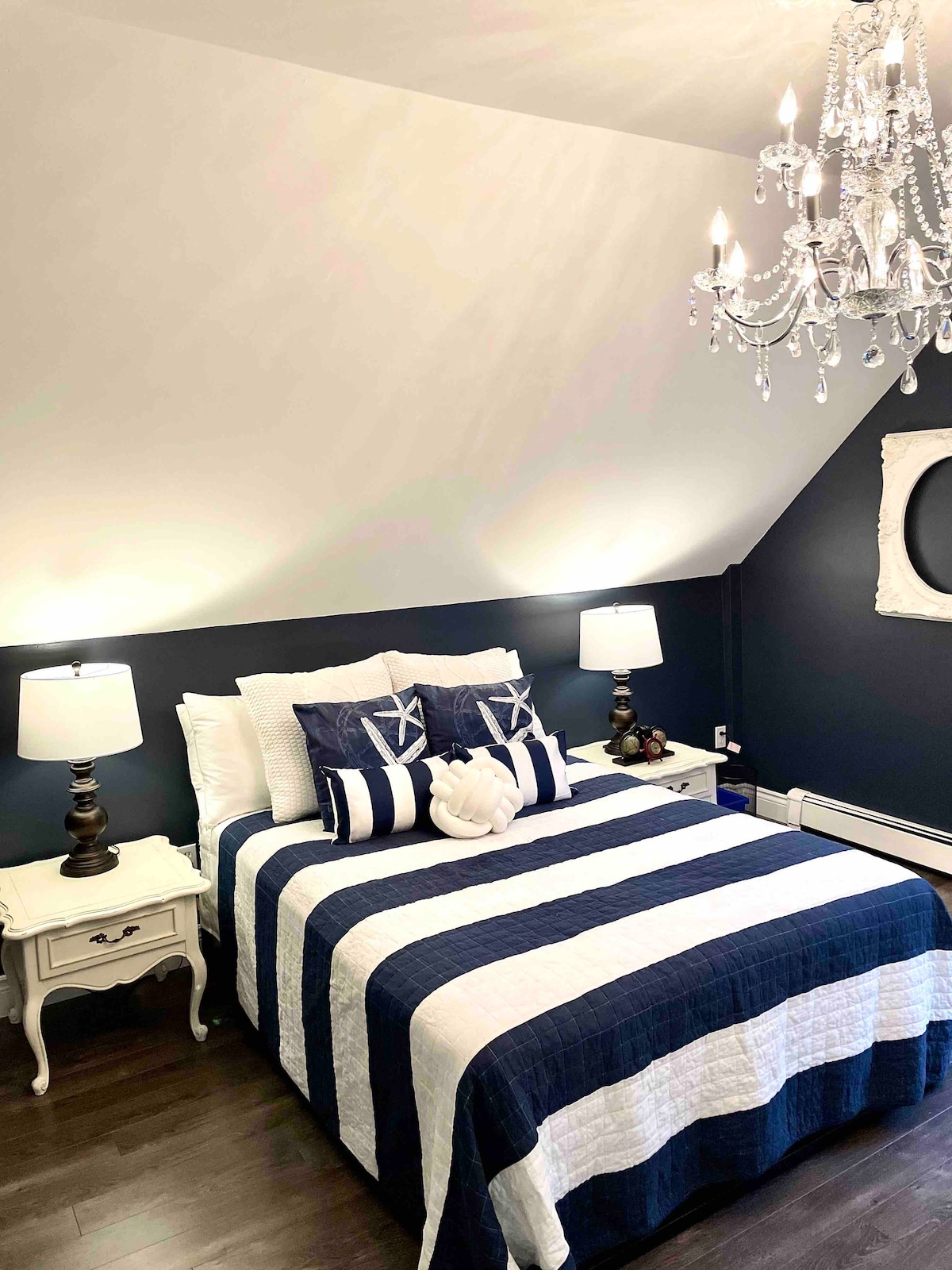 The master bedroom features a queen-sized bed dressed in a navy and white striped quilt, accompanied by decorative pillows. Two bedside tables with lamps illuminate the space, while a chandelier adds elegance. Dark walls and a light ceiling enhance the room's nautical theme.
