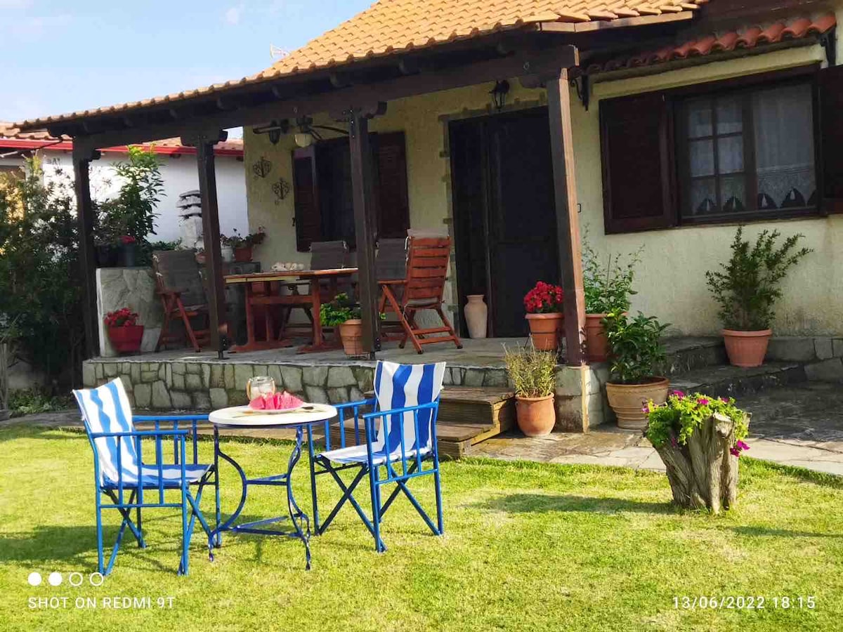 A charming outdoor seating area is set in front of a beach house. Two blue and white striped chairs are positioned around a small round table, accompanied by a potted plant. The porch features wooden furniture and vibrant flowers, creating a welcoming entrance.